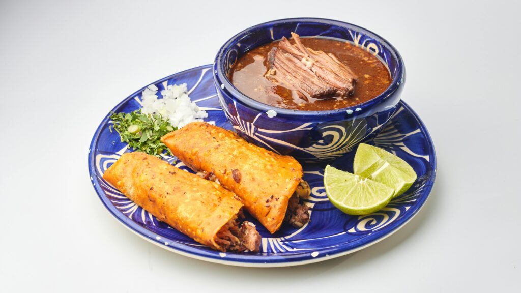 Delicious birria tacos served with rich consommé, onions, herbs, and lime on a colorful plate.