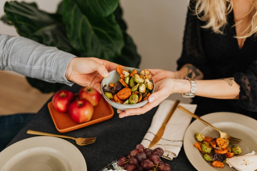Two hands sharing a bowl of colorful roast vegetables on a table with plates, fruits, and cutlery.