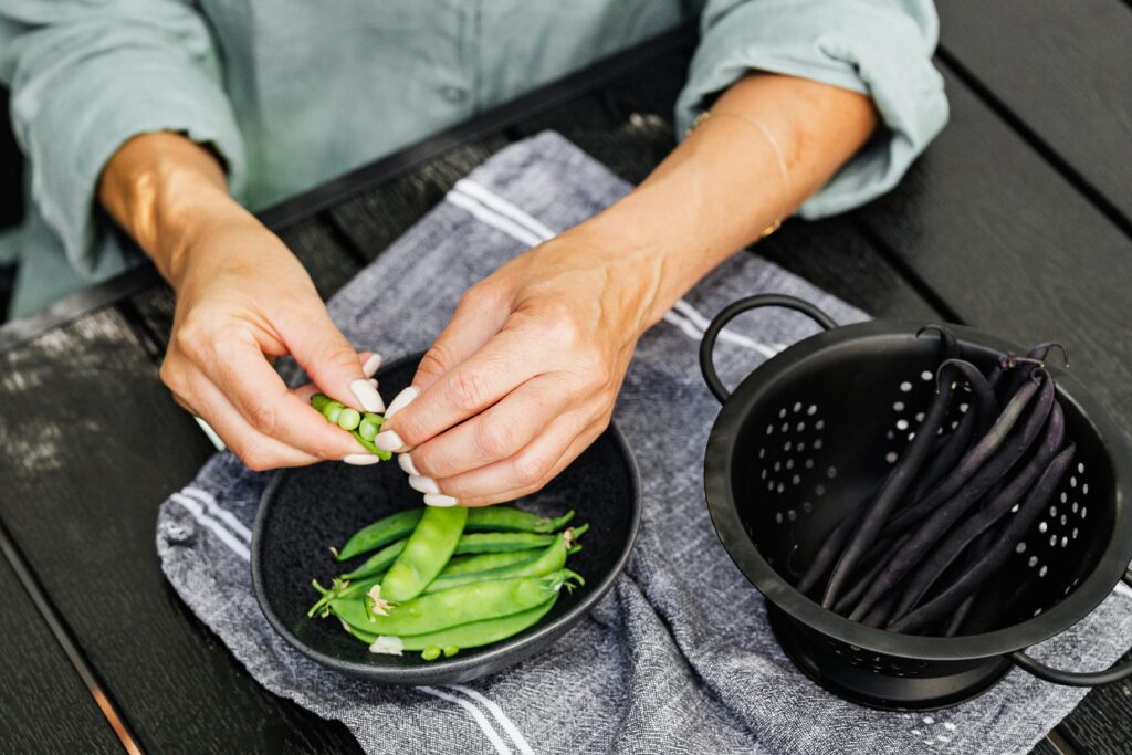 Close-up of hands shelling fresh snap peas over a black bowl outdoors.